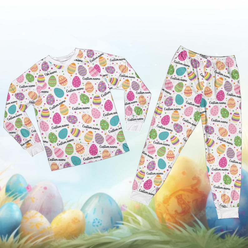 May include: White Easter-themed pajama set with a long-sleeved top and pants. The set is covered in colorful Easter eggs with the text "Custom name" on each egg. The eggs are in various colors, including pink, blue, green, and yellow.