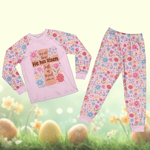 May include: A light pink pajama set featuring a long-sleeved top and pants. The top has a cross design with the text "He has Risen" and floral accents. The sleeves and pants are covered in a pattern of flowers, eggs, and other spring elements.