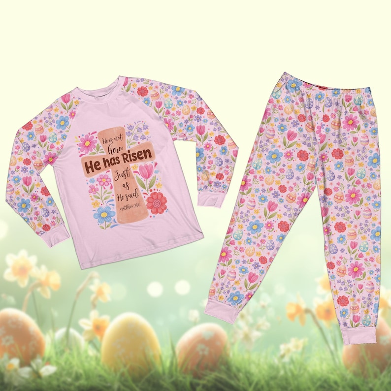 May include: A light pink pajama set featuring a long-sleeved top and pants. The top has a cross design with the text "He has Risen" and floral accents. The sleeves and pants are covered in a pattern of flowers, eggs, and other spring elements.