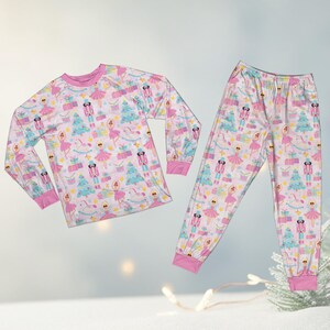 May include: A two-piece pajama set featuring a pink, blue, and white pattern. The long-sleeved top and pants are adorned with images of ballerinas, nutcrackers, Christmas trees, and wrapped gifts. The cuffs and neckline are trimmed in pink.