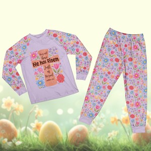 May include: A light purple pajama set featuring a long-sleeved shirt and pants. The shirt has a cross design with the text "He has Risen". The set is decorated with floral and Easter egg patterns.