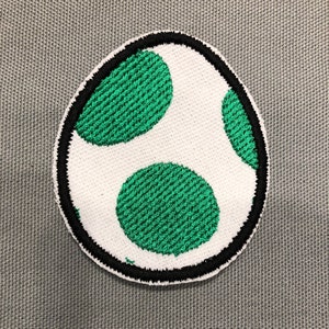 May include: Embroidered patch featuring a white egg shape with three green spots. The egg is outlined in black stitching. The patch is likely for clothing or accessories.