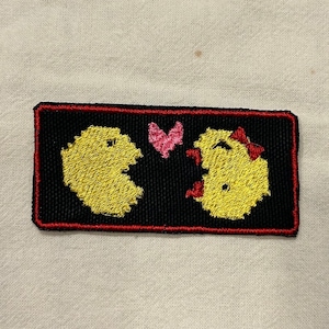 May include: An embroidered patch featuring two yellow Pac-Man characters with a pink heart between them, on a black background with a red border.