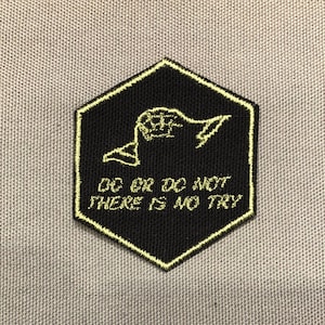 Op de afbeelding: Zwarte zeshoekige patch met een gele rand en de tekst "DO OR DO NOT THERE IS NO TRY". De patch heeft een gele afbeelding van een hand die een lichtzwaard vasthoudt.