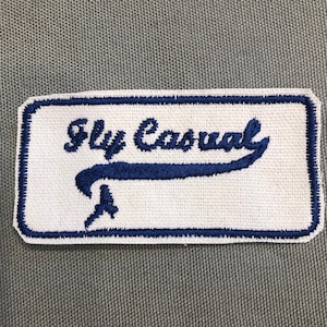 Könnte beinhalten: Blau-weißer gestickter Aufnäher mit dem Text "Fly Casual" in einer kursivschriftlichen Schriftart. Der Aufnäher zeigt ein stilisiertes Flugzeugdesign.