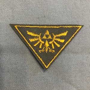 May include: A gray triangular patch with a yellow embroidered design of a stylized bird with wings spread. The design is a popular symbol from the Legend of Zelda video game series.