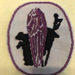 May include: Purple and black embroidered patch with a stylized image of a person standing in front of a large crystal.
