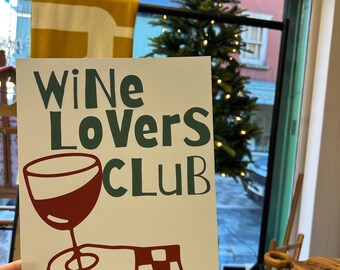 May include: A white sign with the words "Wine Lovers Club" in green and a graphic of a hand holding a wine glass filled with red wine. The text "7pm-late all weekend" is below the graphic. The sign is held in front of a window.