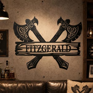 Op de afbeelding: Een zwart metalen wanddecoratie met twee gekruiste bijlen met ingewikkelde decoratieve patronen op de bladen en handvatten. Een horizontale rechthoekige plaat is in het midden van de bijlen geïntegreerd en toont de naam FITZGERALD.