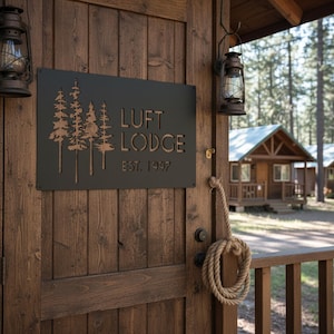 K&ouml;nnte beinhalten: Ein schwarzes Metallschild mit der Aufschrift "LUFT LODGE EST. 1997" und einem Baumdesign ist an einer Holzt&uuml;r befestigt. Zwei Vintage-Laternen h&auml;ngen auf beiden Seiten der T&uuml;r. Ein Seil ist am T&uuml;rgriff befestigt. Die Szene deutet auf eine rustikale Lodge hin.