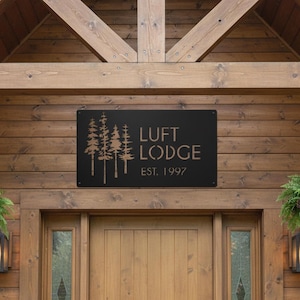 K&ouml;nnte beinhalten: Schwarzes Metallschild mit der Aufschrift "LUFT LODGE EST. 1997" und einer Waldsilhouette. Das Schild ist an einer Holzwand &uuml;ber einer Holzt&uuml;r angebracht. Das Schild hat eine rechteckige Form.