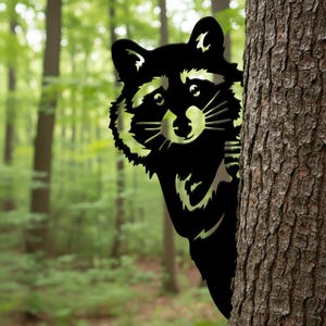 May include: A black metal raccoon peeking from behind a tree trunk. The raccoon has detailed features, including a fluffy face and pointed ears. The background is a blurred forest scene with green foliage.