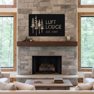K&ouml;nnte beinhalten: Schwarzes Metallschild mit der Aufschrift "LUFT LODGE EST. 1997" und einem Baumdesign. Es ist an einem Steinkamin &uuml;ber einem Holzkamin befestigt. Der Raum hat gro&szlig;e Fenster und eine rustikale &Auml;sthetik.