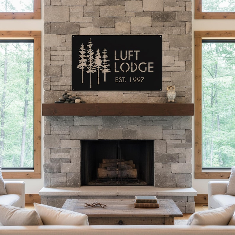 K&ouml;nnte beinhalten: Schwarzes Metallschild mit der Aufschrift "LUFT LODGE EST. 1997" und einem Baumdesign. Es ist an einem Steinkamin &uuml;ber einem Holzkamin befestigt. Der Raum hat gro&szlig;e Fenster und eine rustikale &Auml;sthetik.