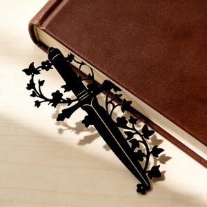 Könnte beinhalten: Schwarzer Metall-Lesezeichen in Schwertform mit floralen und Blatt-Akzenten. Das Lesezeichen steckt in einem braunen, ledergebundenen Buch, das auf einer hellen Holzoberfläche liegt. Das Design ist detailliert.