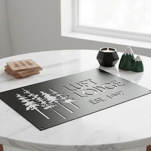 K&ouml;nnte beinhalten: Ein schwarzes Metallschild mit dem Text "LUFT LODGE EST. 1997" und einem ausgeschnittenen Baumdesign. Das Schild steht auf einem wei&szlig;en Marmortisch, zusammen mit Holzuntersetzern, einem schwarzen Kerzenhalter und bergf&ouml;rmiger Dekoration.
