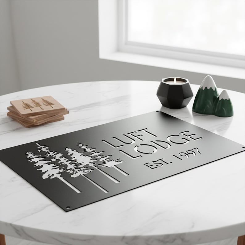 K&ouml;nnte beinhalten: Ein schwarzes Metallschild mit dem Text "LUFT LODGE EST. 1997" und einem ausgeschnittenen Baumdesign. Das Schild steht auf einem wei&szlig;en Marmortisch, zusammen mit Holzuntersetzern, einem schwarzen Kerzenhalter und bergf&ouml;rmiger Dekoration.