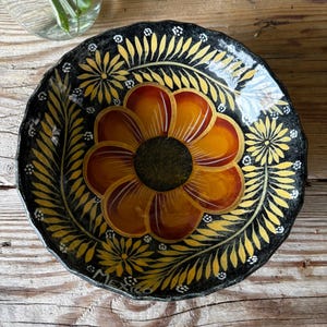 May include: A round, black ceramic bowl with a scalloped edge, featuring a large central orange and yellow flower surrounded by yellow leaf patterns and small white dots. The word MEXICO is painted in white near the bottom rim of the bowl.