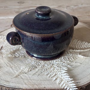 May include: A small, dark blue ceramic casserole dish with a lid and two side handles. The dish has a round shape and sits on a small base. The lid has a small knob on top. The dish is glazed with a glossy finish.