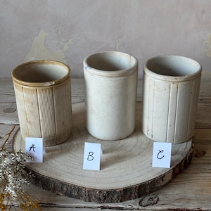 Puede incluir: Tres tarros de cerámica antiguos de diferentes diseños y colores. Los tarros son cilíndricos con la parte superior abierta. Uno es de color beige con estrías verticales, otro es blanquecino y liso, y el tercero es blanquecino con líneas verticales. Están sobre una rodaja de madera.