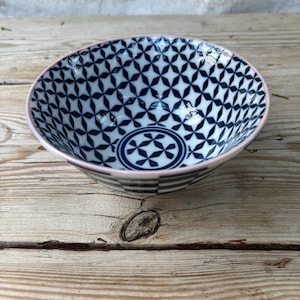 May include: A ceramic bowl with a geometric pattern in navy blue and white. The bowl has a pink rim and a circular design at the bottom. The bowl is on a wooden surface.