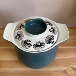 May include: A round, dark teal ceramic casserole dish with two side handles and a matching lid. The lid features a cream-coloured top surface decorated with a repeating pattern of brown and black mushroom illustrations and a dark teal centre knob.