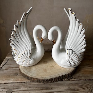May include: A pair of white ceramic swan ornaments with gold detailing on the wings and beaks. The swans are arranged to face each other, forming a heart shape with their necks. They are presented on a wooden slice.