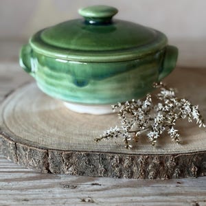 May include: A green ceramic casserole dish with a lid and two handles. The dish is on a wooden slice with dried flowers. The ceramic has a glossy finish and a white base. The dish is approximately 15 cm in diameter.