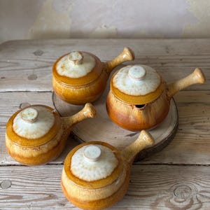 May include: Four ceramic soup bowls with lids and side handles. The bowls have a two-tone design, with cream-coloured lids and amber-coloured bodies. They are arranged on a wooden surface, ideal for serving soup or stew.