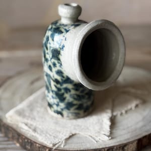 May include: A handcrafted ceramic vase with an unusual, angled form. The vase has a speckled blue and white pattern on its body and a wide, open spout. It is displayed on a wooden slice with a linen cloth.