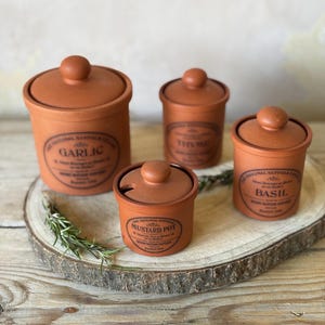 May include: Four terracotta storage jars with lids, labelled Garlic, Thyme, Basil, and Mustard Pot. Each jar features black printed text including The Original Suffolk Collection and Henry Watson Potteries. The mustard pot lid has a small notch.