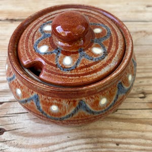 May include: A round ceramic sugar bowl with a lid and a small spoon slot. The bowl is brown with blue and white decorative patterns. The lid has a rounded knob and matching patterns.