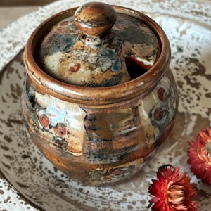 May include: A handmade ceramic sugar bowl with a lid and a small spoon opening. The bowl has a brown, tan, and blue mottled glaze with red accents. It sits on a white, textured plate, with red flowers.