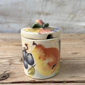 Puede incluir: Un tarro de cerámica con tapa, decorado con diseños de frutas pintados a mano. El tarro cilíndrico tiene una tapa con un pequeño asa y una ranura para una cuchara. Las ilustraciones de frutas incluyen peras, uvas y hojas, en tonos amarillos, marrones y azules.