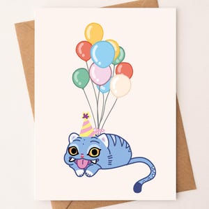 May include: A greeting card featuring a cartoon blue cat with yellow eyes, a party hat, and a long tongue. The cat is holding a bunch of colorful balloons. The card is on a cream background with a brown envelope.