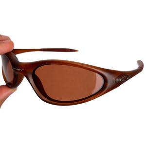 May include: Brown oval-shaped sunglasses with brown lenses. The sunglasses have a curved frame and are held by a hand. The Oakley logo is visible on the frame.