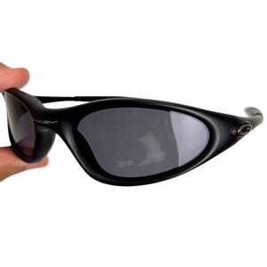 May include: Black sunglasses with a curved, wraparound design. The lenses are dark gray, and the frames are matte black. The Oakley logo is visible on the temple. The sunglasses are held by a hand against a white background.