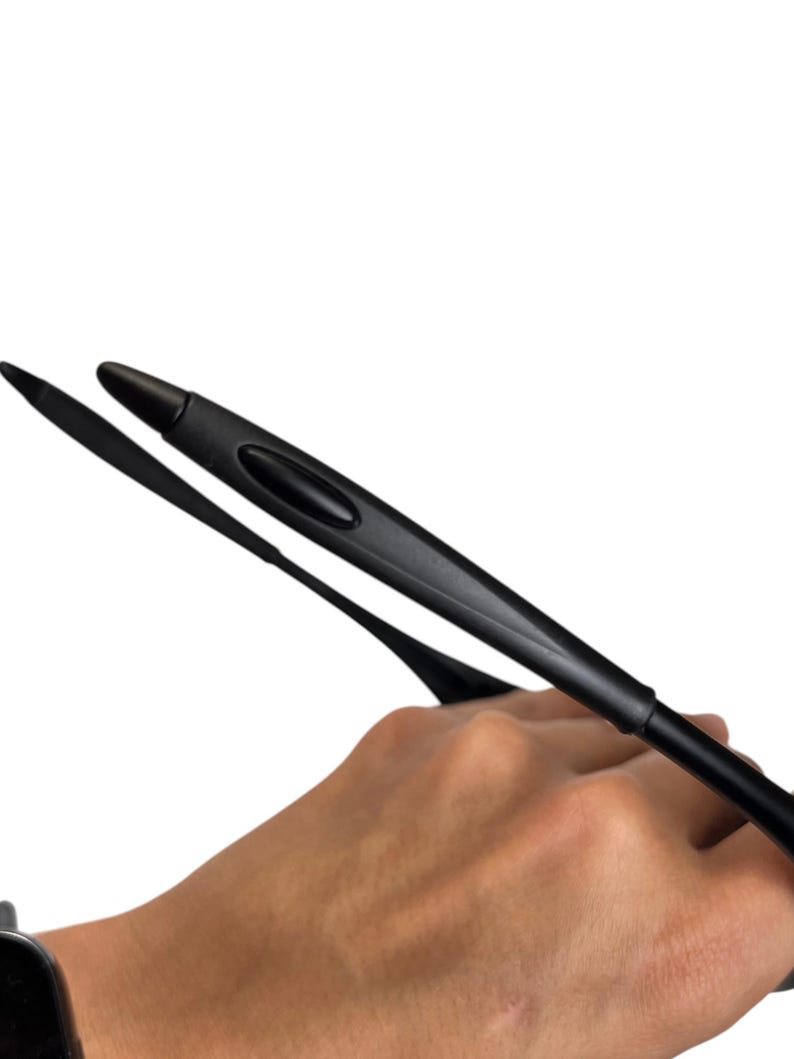 May include: Black eyeglasses arm with a curved design. The arm is held in a hand against a white background. The arm has a rounded tip and a groove along its length.