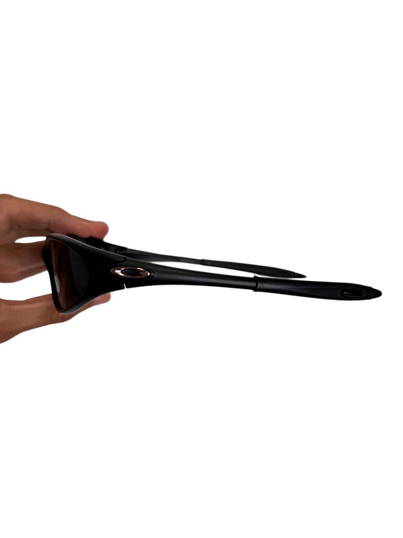May include: Black sunglasses with a sleek, modern design. The sunglasses have dark lenses and a black frame. The arms of the sunglasses are thin and curve slightly. The Oakley logo is visible on the frame.