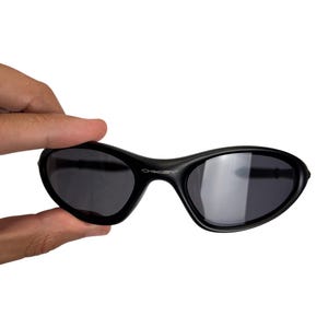 May include: Black sunglasses with a curved frame and dark lenses. The sunglasses are held by a hand against a white background. The brand name "Oakley" is visible on the frame.