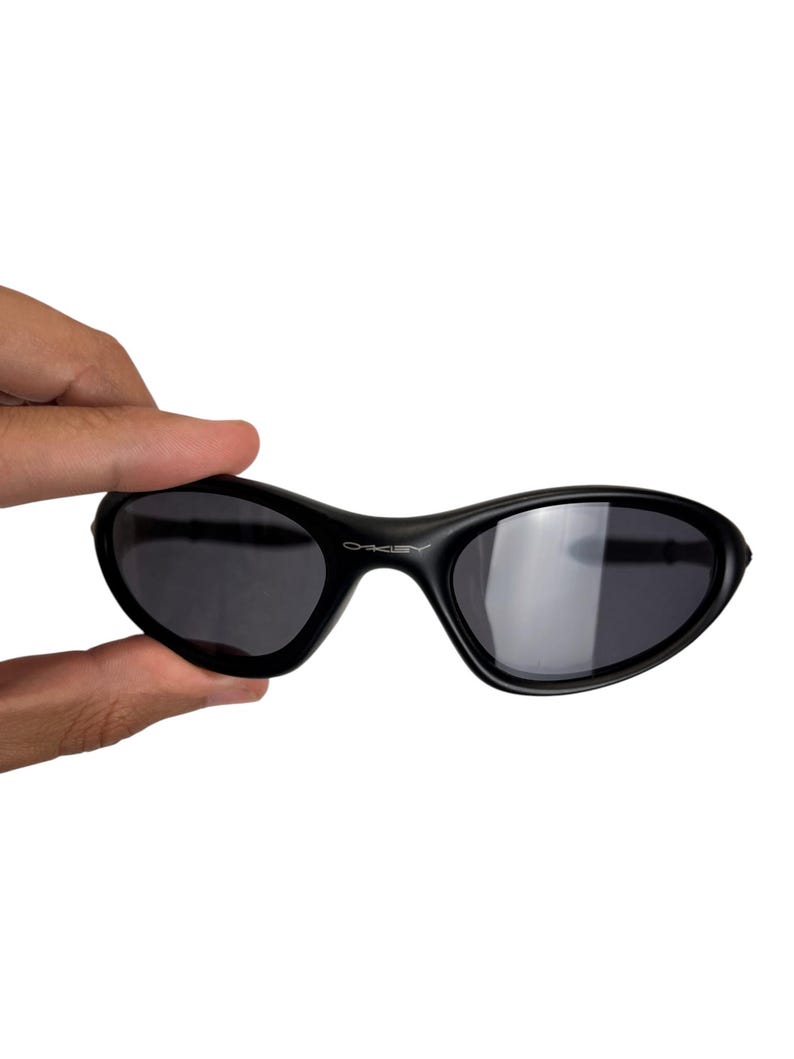 May include: Black sunglasses with a curved frame and dark lenses. The sunglasses are held by a hand against a white background. The brand name "Oakley" is visible on the frame.