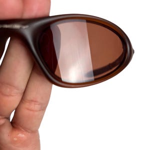 May include: Brown sunglasses with a rounded frame and tinted lenses. The sunglasses are held in a hand, showcasing the design and color of the eyewear. The lenses have a reflective quality.