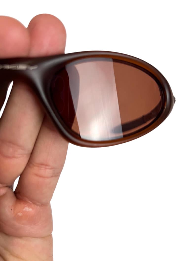 May include: Brown sunglasses with a rounded frame and tinted lenses. The sunglasses are held in a hand, showcasing the design and color of the eyewear. The lenses have a reflective quality.