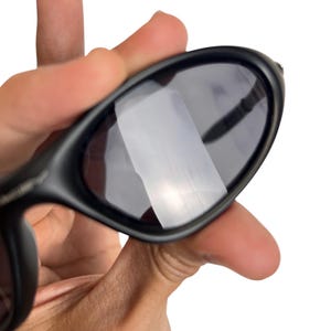 May include: Black sunglasses with dark lenses. The sunglasses have a sleek, curved design and are held in a hand against a white background. The lenses reflect light.