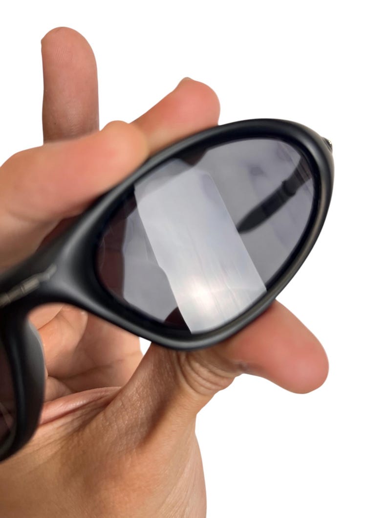 May include: Black sunglasses with dark lenses. The sunglasses have a sleek, curved design and are held in a hand against a white background. The lenses reflect light.