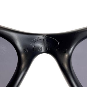 May include: Close-up of black sunglasses with gray lenses. The brand name "MINUTE" is printed in white on the frame. The sunglasses have a curved design and a sporty aesthetic.