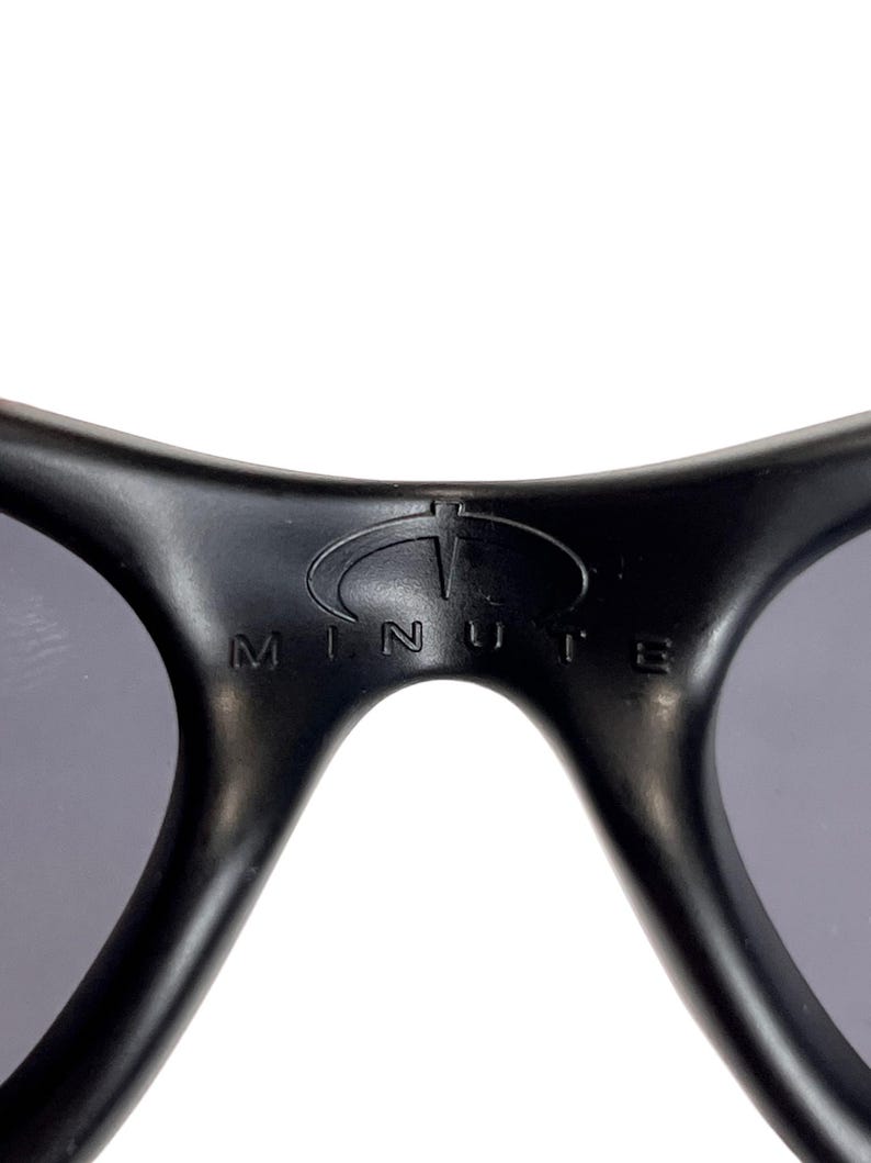 May include: Close-up of black sunglasses with gray lenses. The brand name "MINUTE" is printed in white on the frame. The sunglasses have a curved design and a sporty aesthetic.