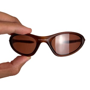May include: Brown Oakley sunglasses with a curved frame and brown lenses. The Oakley logo is visible on the frame. The sunglasses are held by a hand against a white background.