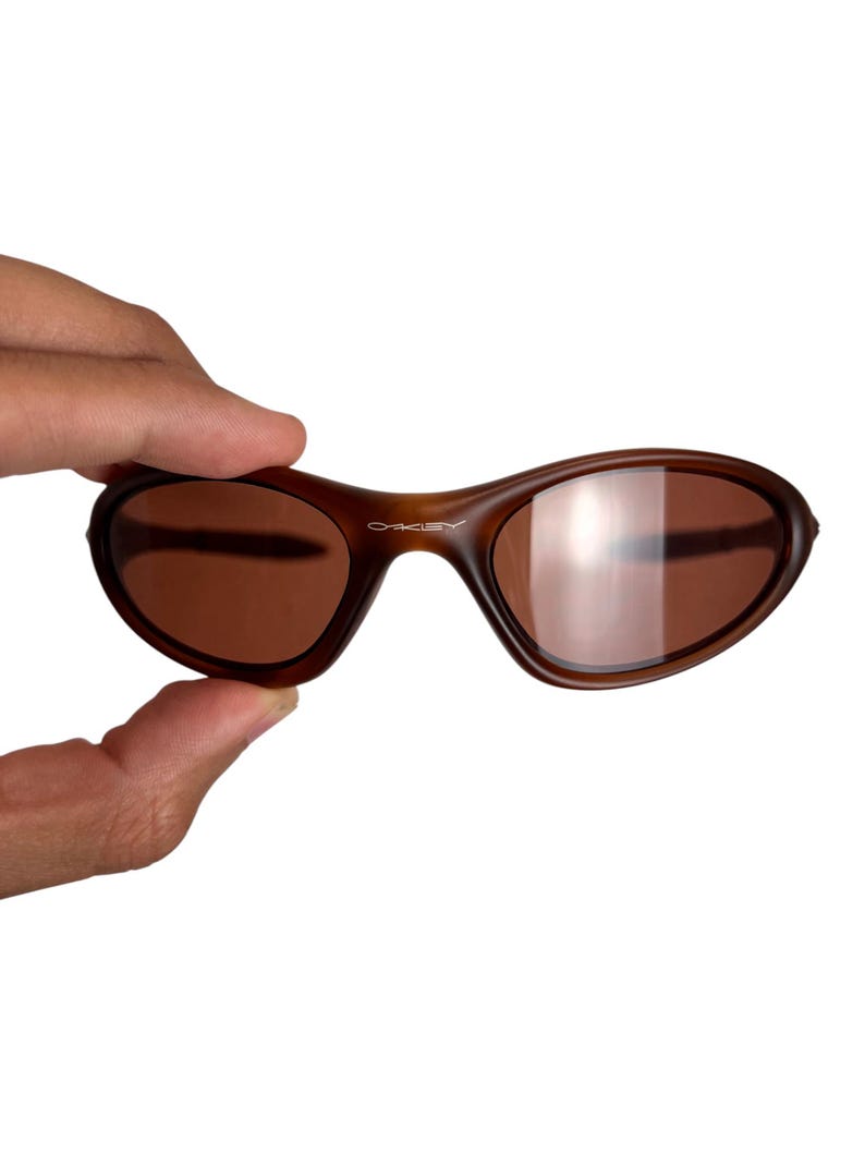 May include: Brown Oakley sunglasses with a curved frame and brown lenses. The Oakley logo is visible on the frame. The sunglasses are held by a hand against a white background.