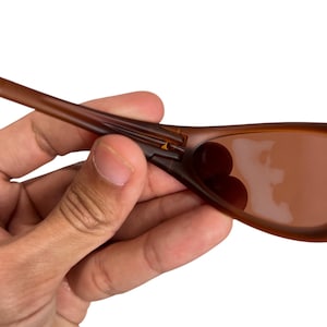 May include: Brown sunglasses with a curved frame and tinted lenses. The sunglasses are held by a hand, showcasing the frame's design and lens color. The lenses appear to be a darker shade of brown.